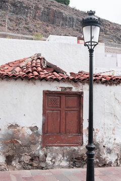 A weathered white cottage with crumbling plaster and a terracotta tile roof features a rustic wooden window. 