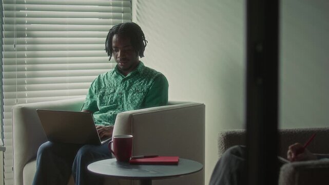 Young African American businessman with dreadlocks sitting in comfortable armchair, using his laptop next to coffee table in modern office workspace