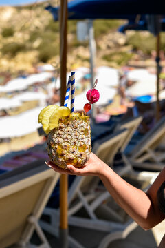 A person holds a tropical cocktail served inside a hollowed pineapple shell at a beach resort. The drink is garnished with fresh fruit slices, straws, and a decorative pick.