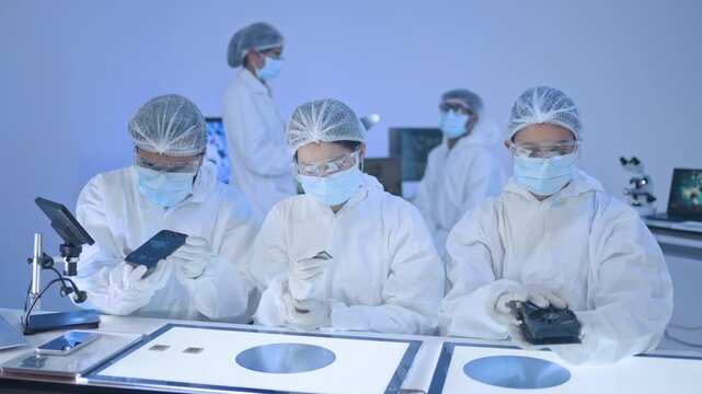 Three specialists in white coveralls and masks checking devices over illuminated table with wafer trays, semiconductor production facility.