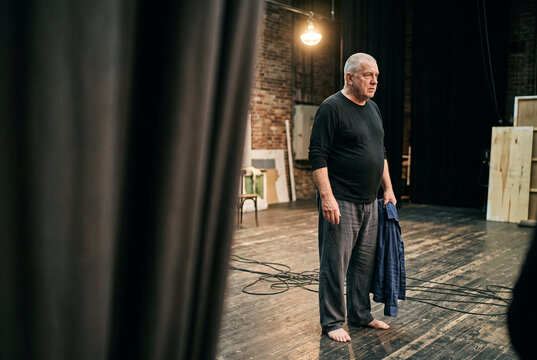 Elderly actor standing barefoot on a stage during a theater rehearsal in a rustic venue for performance background and drama poster