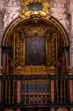 Vertical view looking into an ornate side chapel featuring a gilded baroque altarpiece with a central painting framed by dark marble arches, gold cartouche inscription above, and a dark wooden and mar