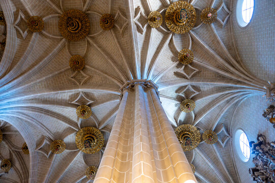 Low angle view looking directly up along a tall stone pillar toward the intricate gothic star-ribbed vault ceiling adorned with golden decorative bosses (claves de b&oacute;veda) and small circular windows i