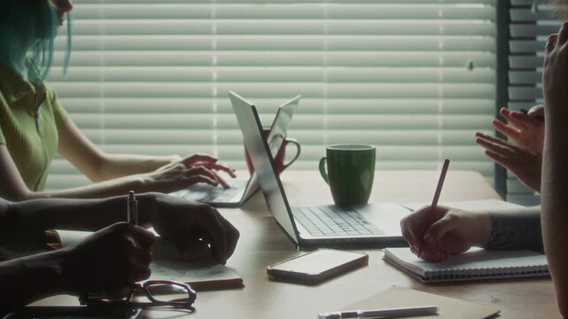 Close up shot of hands of diverse group of colleagues sitting around wooden table, typing on laptop, writing in notebooks and discussing business project during office meeting