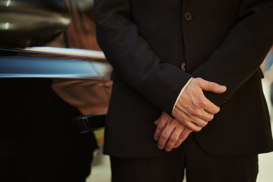 Close up of middle aged male driver standing beside luxury car, clasping hands in formal service pose. Ideal for chauffeur, executive transport, security, premium service marketing