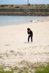 Child explores sandy beach near calm water. Curious posture suggests joyful discovery. Background shows gentle waves and distant breakwall. Ideal for parenting, childhood, or nature themes © Masakazu Tokashiki