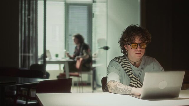 Focused gen Z businesswoman with arm tattoos and yellow glasses typing on laptop at desk in dark office in evening, with colleague working in background
