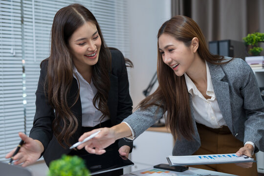 Two professional women work together at a desk, reviewing information on a laptop, collaborating on teamwork and business planning, and analyzing data.