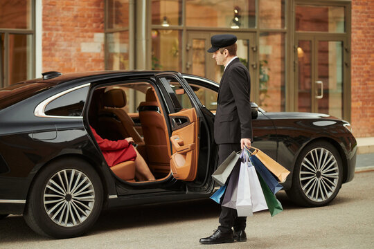 Caucasian chauffeur standing by luxury sedan, holding shopping bags while female client entering rear seat outside hotel, representing premium retail service. Side view for travel marketing
