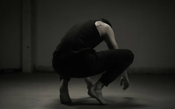 A person in a squatting yoga or stretching pose with hands reaching toward the floor in a dimly lit room