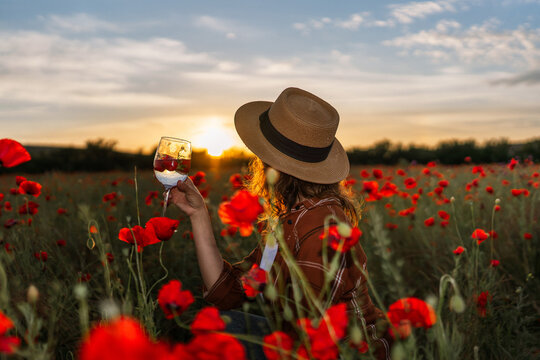 Golden hour sunset features woman wearing straw hat holding wine glass in field of Papaver rhoeas