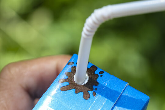 Hand holding small blue drink carton with white bendy straw inserted and chocolate splash stain, close up, outdoor natural light