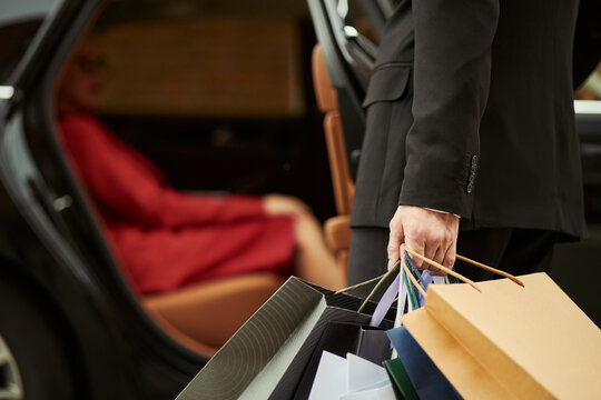 Middle aged chauffeur carrying shopping bags beside luxury car while female client waiting inside, suggesting premium retail service. Ideal for concierge, personal shopping, luxury transport marketing