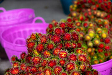 Naklejka premium Rambutan fruit pile with vibrant red spiky skins and green hair displayed in purple plastic baskets at market, colorful tropical produce and fresh harvest evoke lively atmosphere