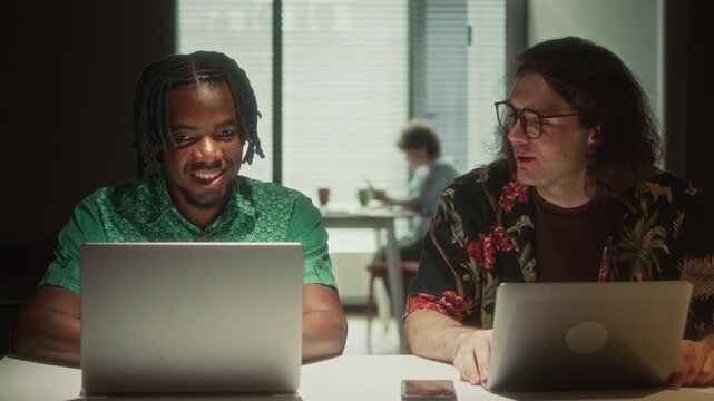 Young African American male programmer and his Caucasian coworker discussing data on laptop screen in dark office while working overtime in evening