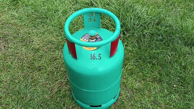 A green 16.5 kg LPG gas cylinder placed upright on a grassy field outdoors. The steel liquefied petroleum gas cylinder features a protective ring around the valve . in malaysia