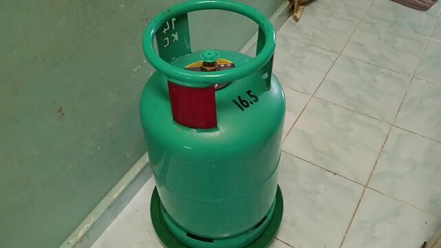 A green 16.5 kg LPG gas cylinder standing upright on a tiled indoor floor against a painted wall. The household cooking gas cylinder features a protective top guard . photo taken in malaysia