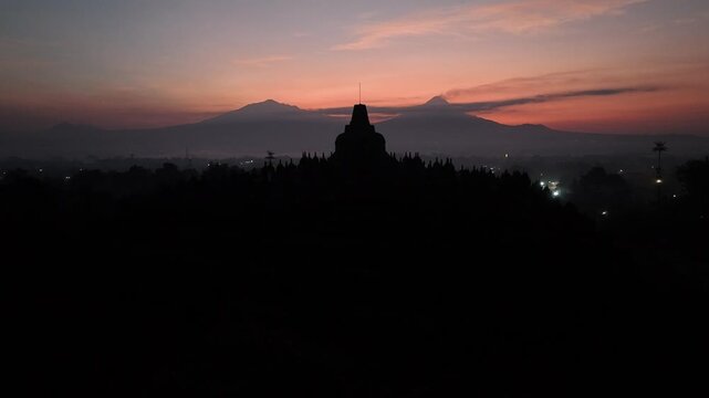 Aerial footage of the silhouette of Borobudur Temple at sunrise, with Mount Merapi and Mount Merbabu in the background.