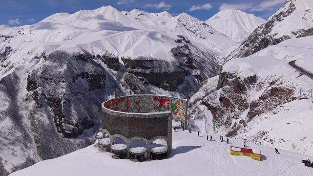 Drone orbit around the Georgia-Russia Friendship Monument in Gudauri, revealing snowy mountain terrain under bright winter sunlight