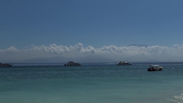 A serene beach scene with clear blue water and distant boats. The sky is bright with a few clouds, creating a peaceful atmosphere.