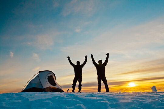 Two friends raise their arms as sun sets on snowy campsite