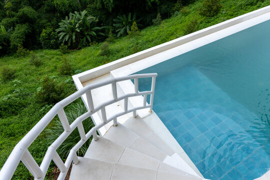 Infinity pool staircase with white railing overlooking lush green hillside, tranquil blue water and modern tiled edge creating serene tropical retreat