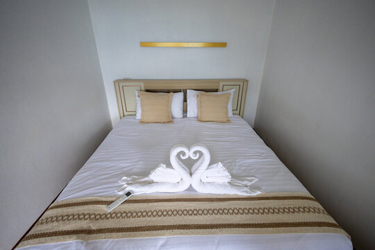 White hotel bed with two towel swans forming heart, beige pillows and decorative runner creating romantic welcome atmosphere