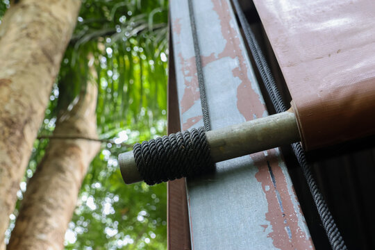 Close up of metal rod wrapped with black braided rope on weathered metal frame beside tropical trees, showing texture and outdoor workshop detail