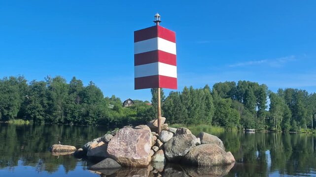 Red and white striped navigation beacon on rocks by the river under blue sky, orientation symbol, travel route, safe navigation, summer day.