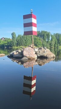 Striped navigation beacon reflected in still river water with rocky islet and trees, river landscape, wayfinding symbol, travel route, calm morning.