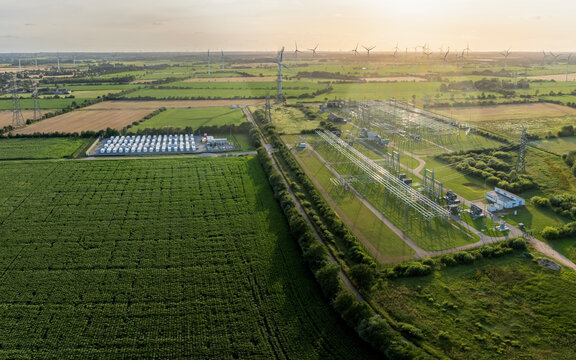 Renewable energy hub with with high-voltage electrical substation, scale battery energy storage system (BESS) composed of white container units, and wind turbines lined horizon.  