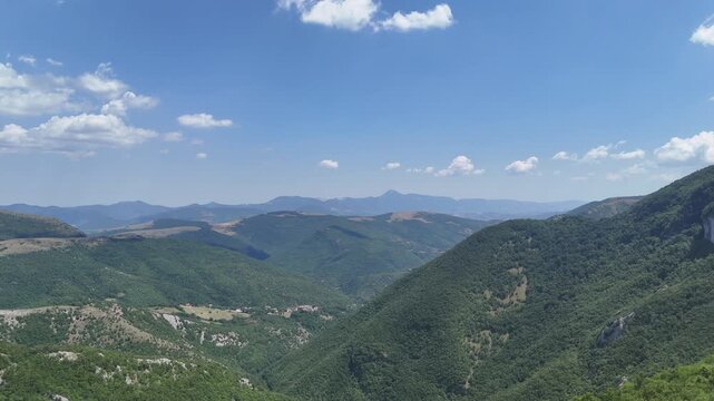 Ampio paesaggio montano con colline verdi, vallate ricoperte di vegetazione fitta e montagne lontane dai toni azzurri, sotto un cielo limpido con nuvole bianche sparse