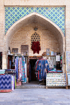 Souvenir shop inside a pointed brick arch of Nadir Divan Begi Madrasah in Bukhara, Uzbekistan. Colorful textiles, scarves, and magnets displayed beneath mosaic tilework. Ideal for travel.