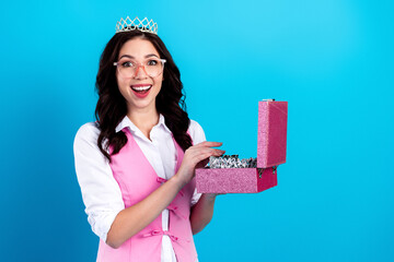 Surprised young woman wearing a tiara holding an open pink jewelry box filled with sparkling jewels against a bright blue background © deagreez