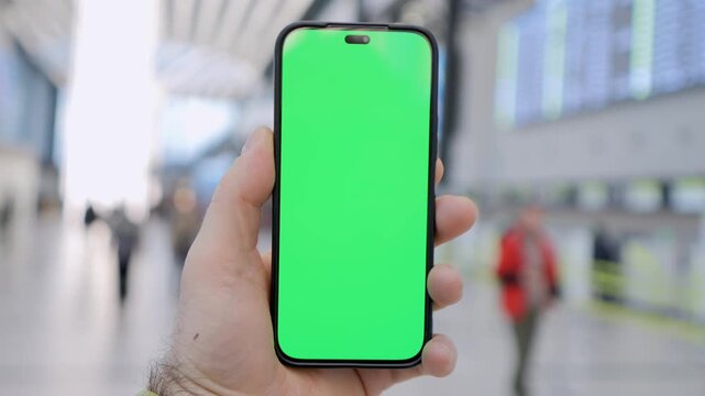 Hand holds phone with green screen. Background shows busy train station. Focus on device for app or ad mockups. Realistic human grip adds authenticity. Useful for travel, tech