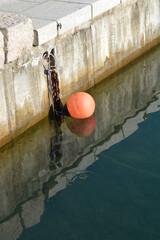 Fototapeta premium A vertical shot of a bright orange mooring buoy floating on calm sea water next to a concrete and stone dock wall with water reflection