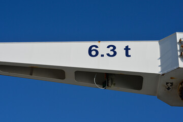 A close up shot of a white industrial metal crane arm showing a black weight load capacity sign text reading six point three tons against a blue sky © nahhan