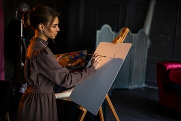 Naklejka premium Young woman painting at easel in dim studio with dramatic shadows and focused expression