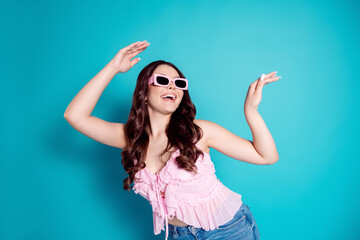 Fototapeta na wymiar Joyful young woman wearing pink sunglasses and ruffled top dancing against a vibrant blue background, smiling carefree and fashionable in casual denim