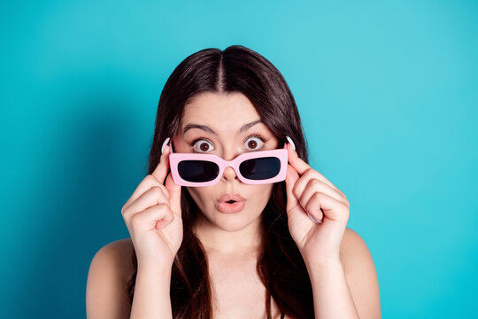Surprised young woman holding pink sunglasses down over eyes against bright blue background, playful summer portrait with wide-eyed expression