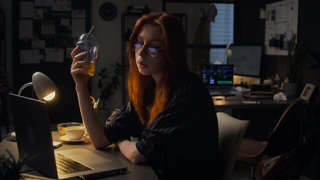 Caucasian young adult woman wearing glasses sitting at office desk at night, holding drink and typing on laptop. Clip is suitable for themes of overtime work, burnout, or freelance projects
