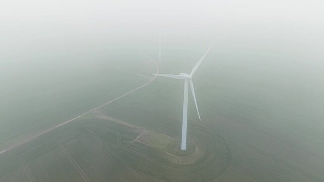 Aerial reveal shot rising through a dense fog layer to discover giant wind turbines spinning at Cotton Farm wind joint, Cambridgeshire, UK.