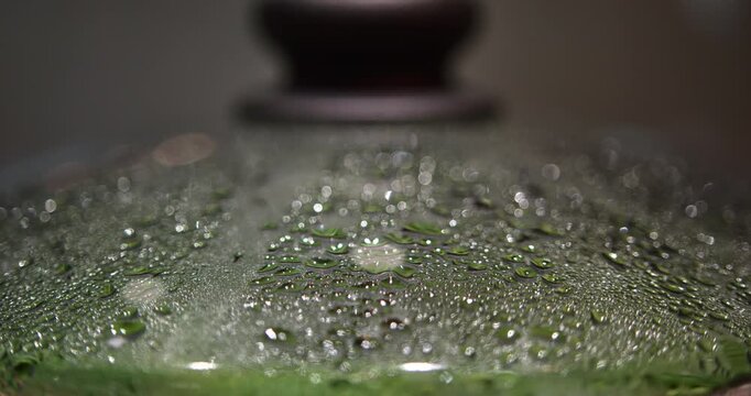 Tight macro view emphasizes large water drops merging on a transparent glass cooking lid while bright green beans remain visible below through steam and shifting reflections