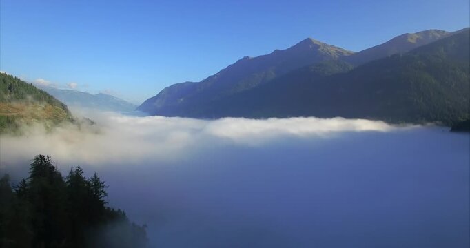 Smooth sideward drone flight follows dense fog spread across an alpine valley in Austria with a green forest edge dark mountain ridge distant peaks and cloudless blue sky