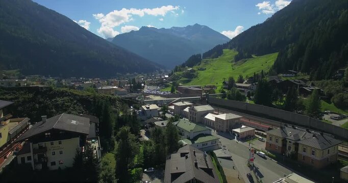 Forward aerial glide crosses Bad Gastein railway station with parallel tracks moving cars hotel buildings and a bright green ski slope leading toward rugged mountain peaks