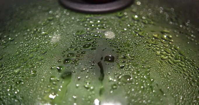 Top down macro view studies rounded condensation droplets and wet streaks on a glass pan lid with green beans visible beneath and a dark knob edging into the shallow focus frame