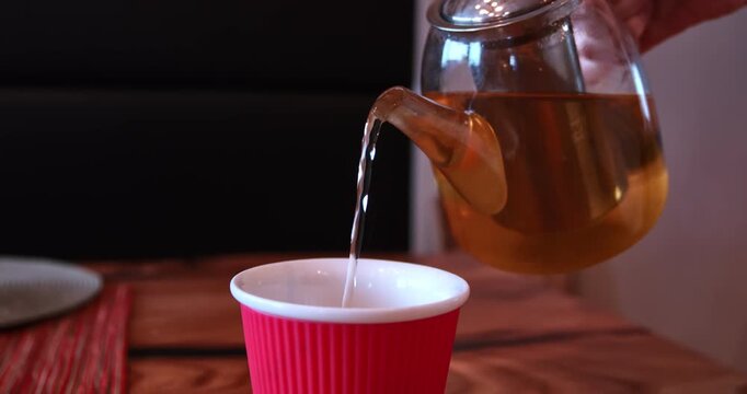 Static close view shows a hand tilting a transparent glass teapot and pouring a steady stream of warm amber tea into a bright red cup arranged on a rustic wooden table at home