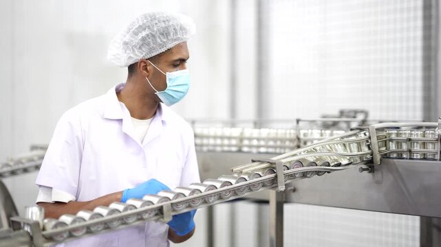 Quality control inspector checking can conveyor line and recording inspection results on tablet in food factory. Quality inspection, food safety, production process, manufacturing, factory operations.