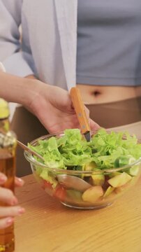 Close up of hands pouring olive oil over a fresh healthy salad in a glass bowl and mixing it on a wooden table.