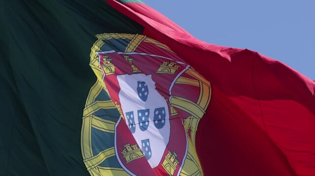 Close-up slow motion of the Portuguese flag swaying gracefully in the wind, vivid red and green fabric illuminated by bright natural sunlight, set against a cloudless blue sky.